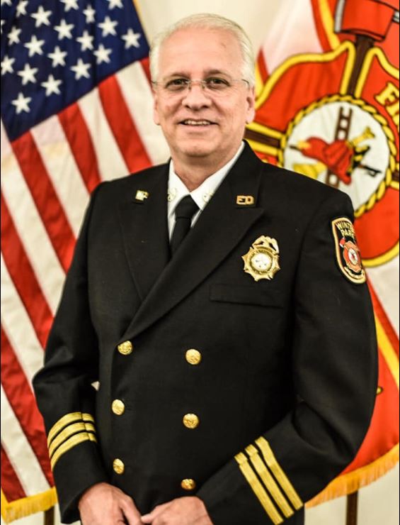 Chief Wales posing in class A uniform in front of USA flag and FD flag